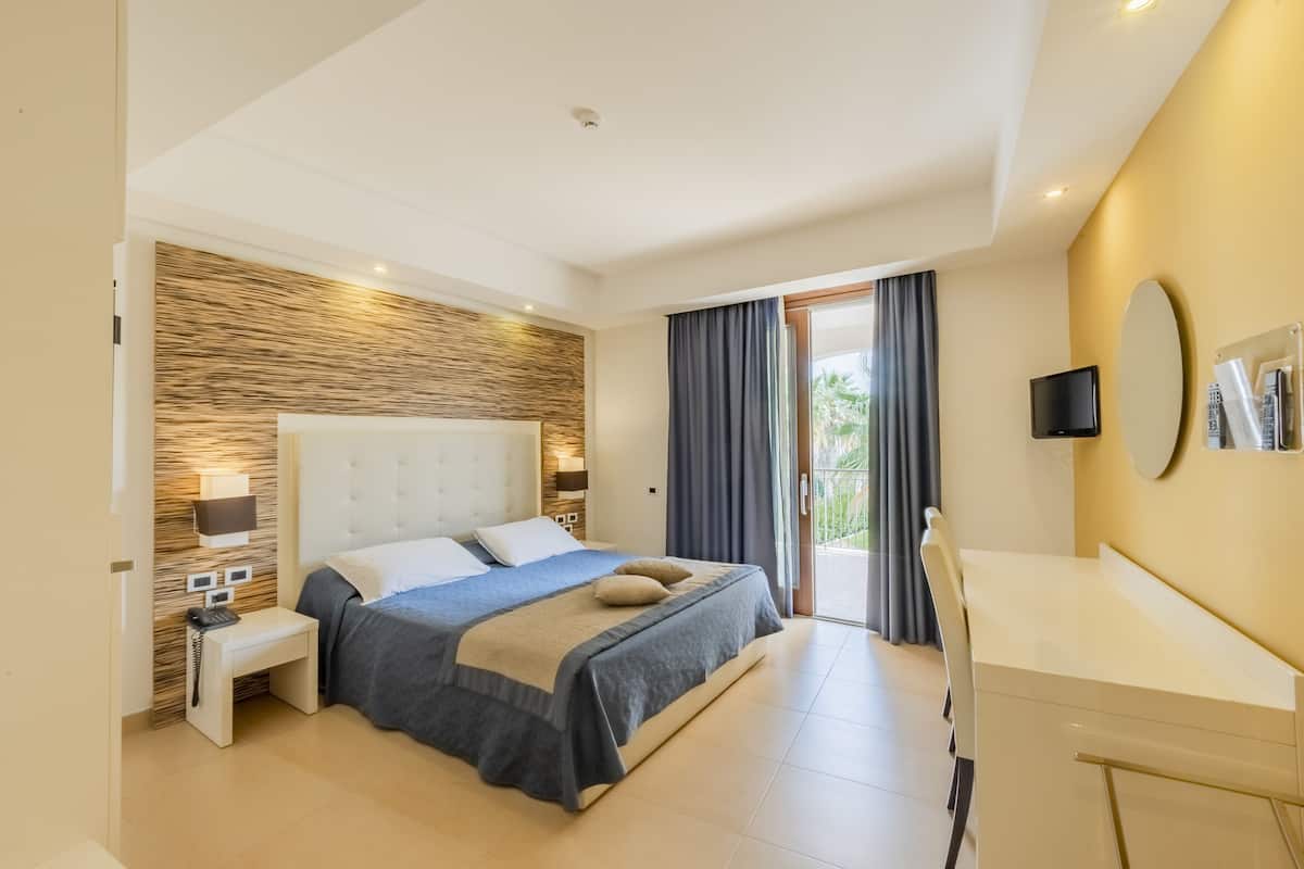 A modern hotel room with a large bed, bedside table, lamp, and a desk with a chair.