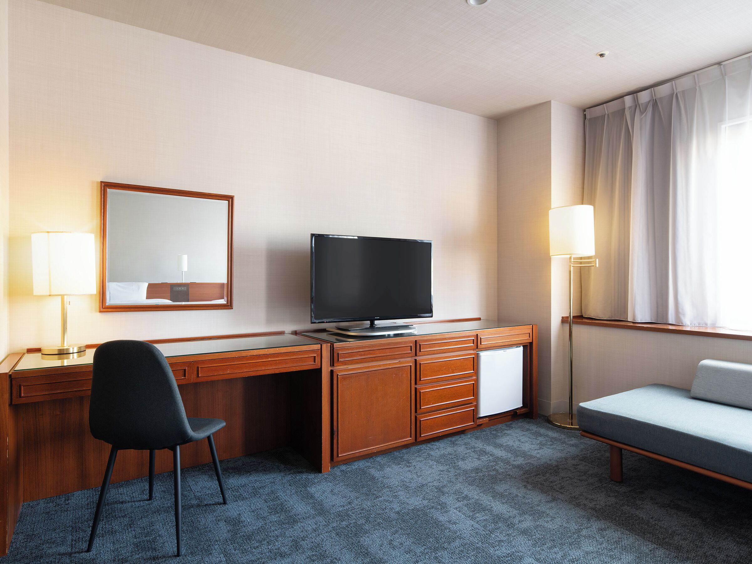 A hotel room with a wooden desk, a television, a chair, a bed, and a window with curtains.