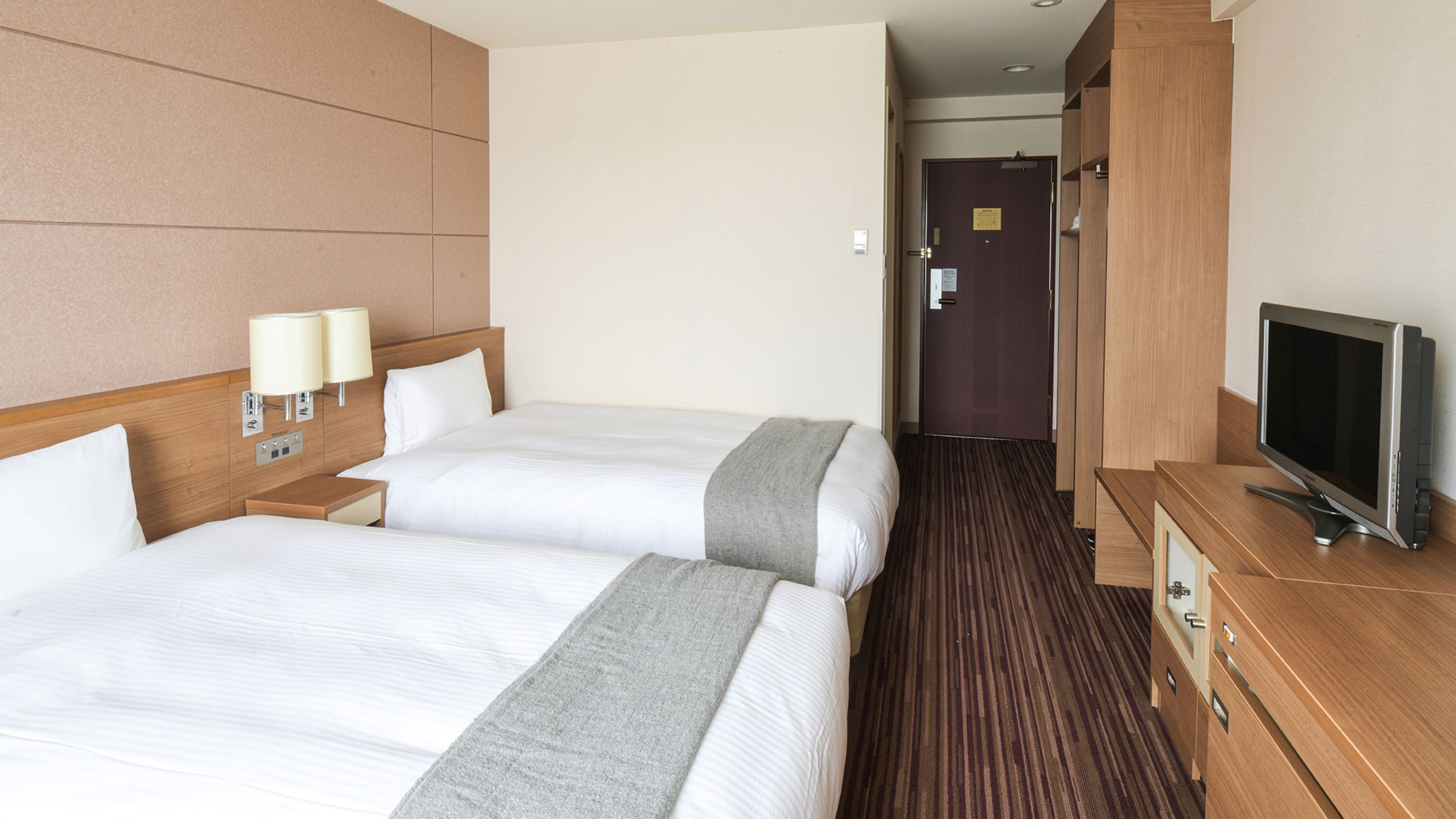 A hotel room with two beds, a TV, a wooden dresser, and a hallway leading to a door.