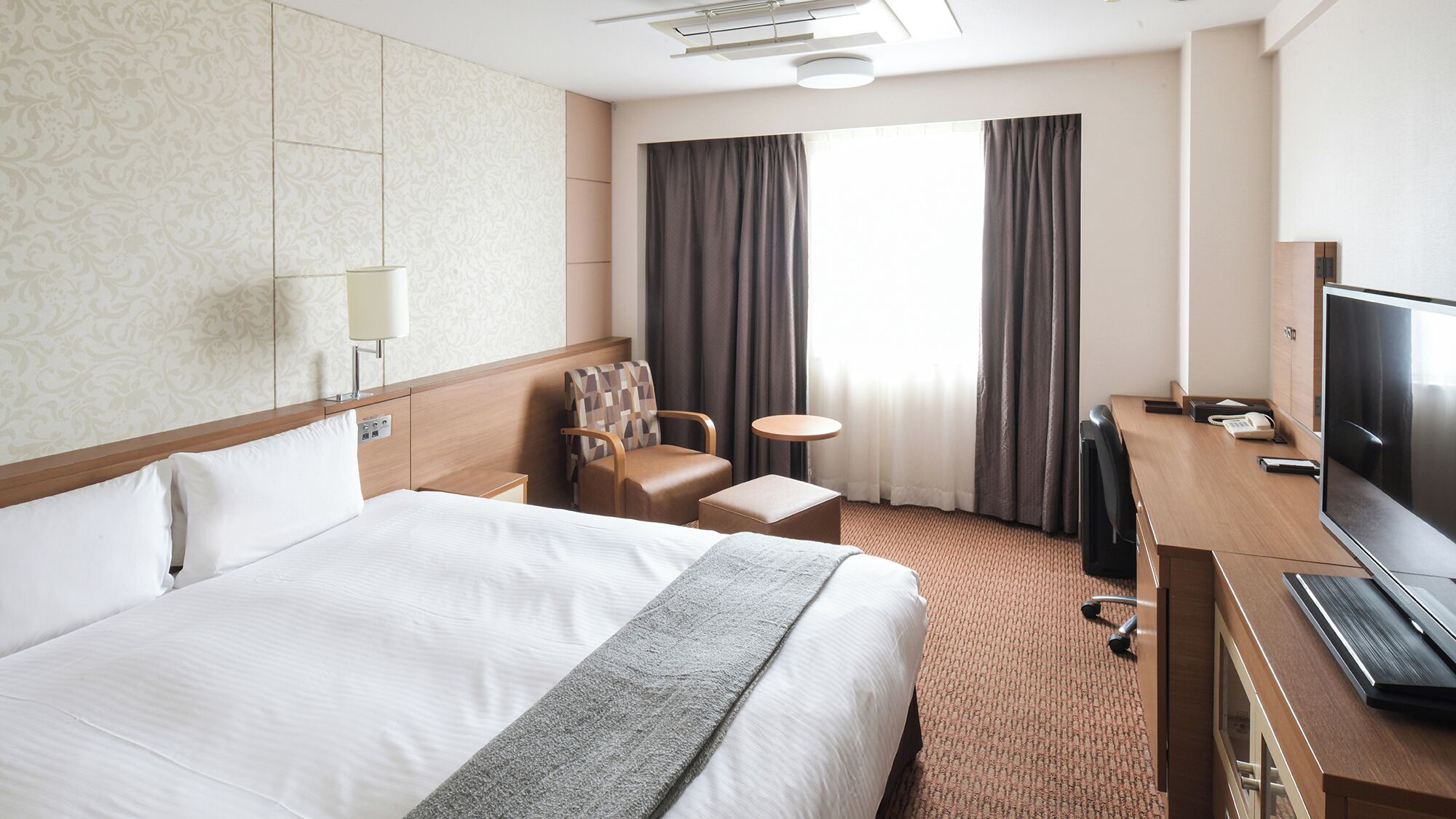 A hotel room with a large bed, a desk with a TV, a chair, a small table, and a window with curtains.