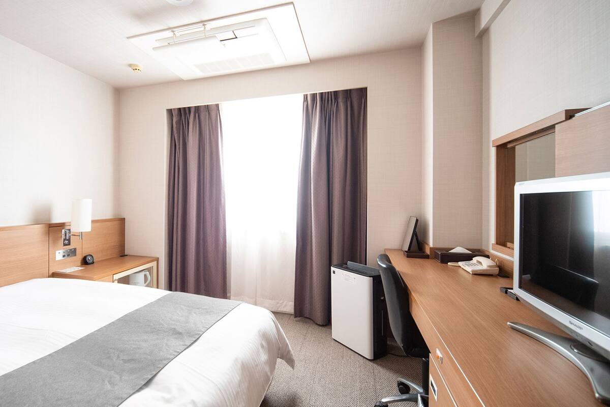 Ein Hotelzimmer mit einem Bett, einem Schreibtisch mit Computer, einem Fernseher und einem Fenster mit Vorhängen.