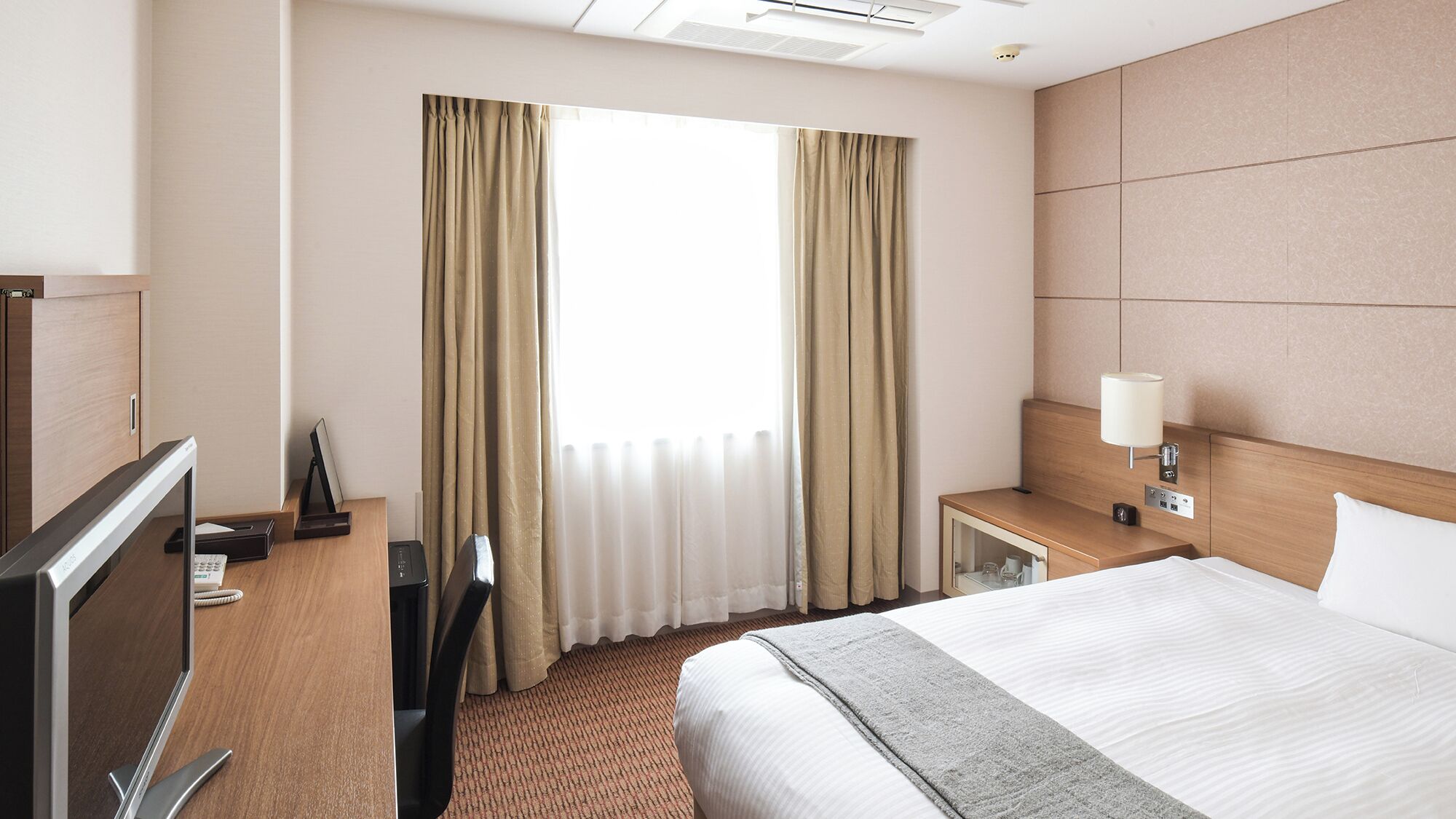 A hotel room with a bed, a desk with a computer, a TV, and a window with curtains.