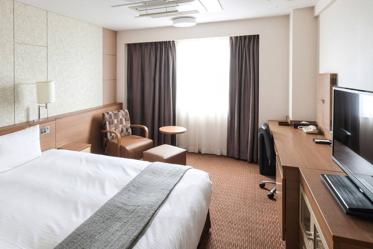 Ein Hotelzimmer mit einem großen Bett, einem Schreibtisch mit Fernseher, einem Sessel, einem kleinen Tisch und einem Fenster mit Vorhängen.