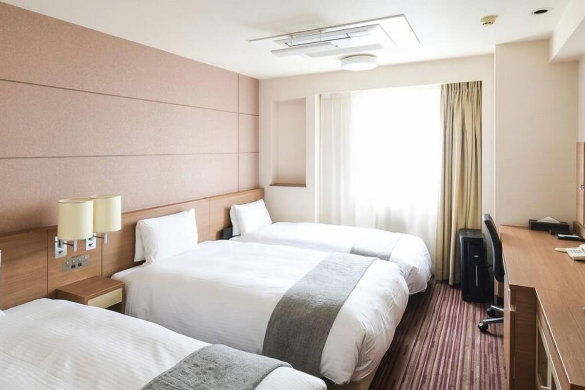 Ein Hotelzimmer mit zwei Betten, einem Schreibtisch, einem Fernseher und einem Fenster mit Vorhängen.