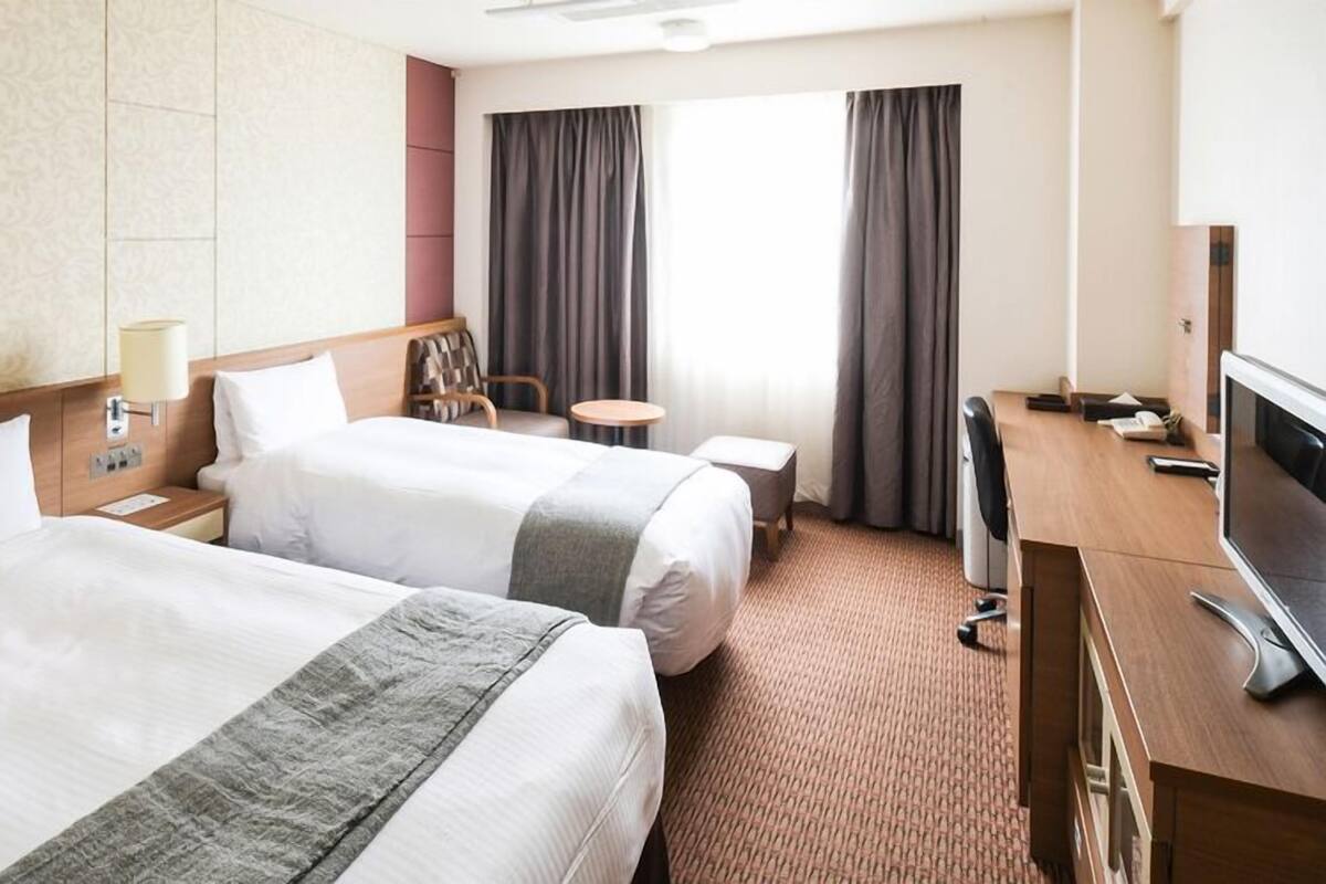 Ein Hotelzimmer mit zwei Betten, einem Schreibtisch mit Fernseher, einem kleinen Tisch und einem Fenster mit Vorhängen.