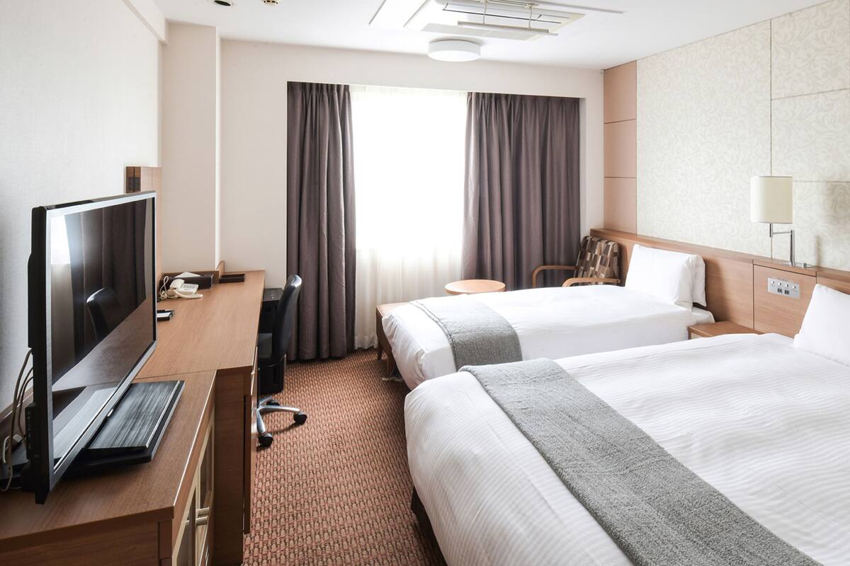 Ein Hotelzimmer mit zwei Betten, einem Schreibtisch mit Fernseher, einem Sessel, einer Lampe und einem Fenster mit Vorhängen.