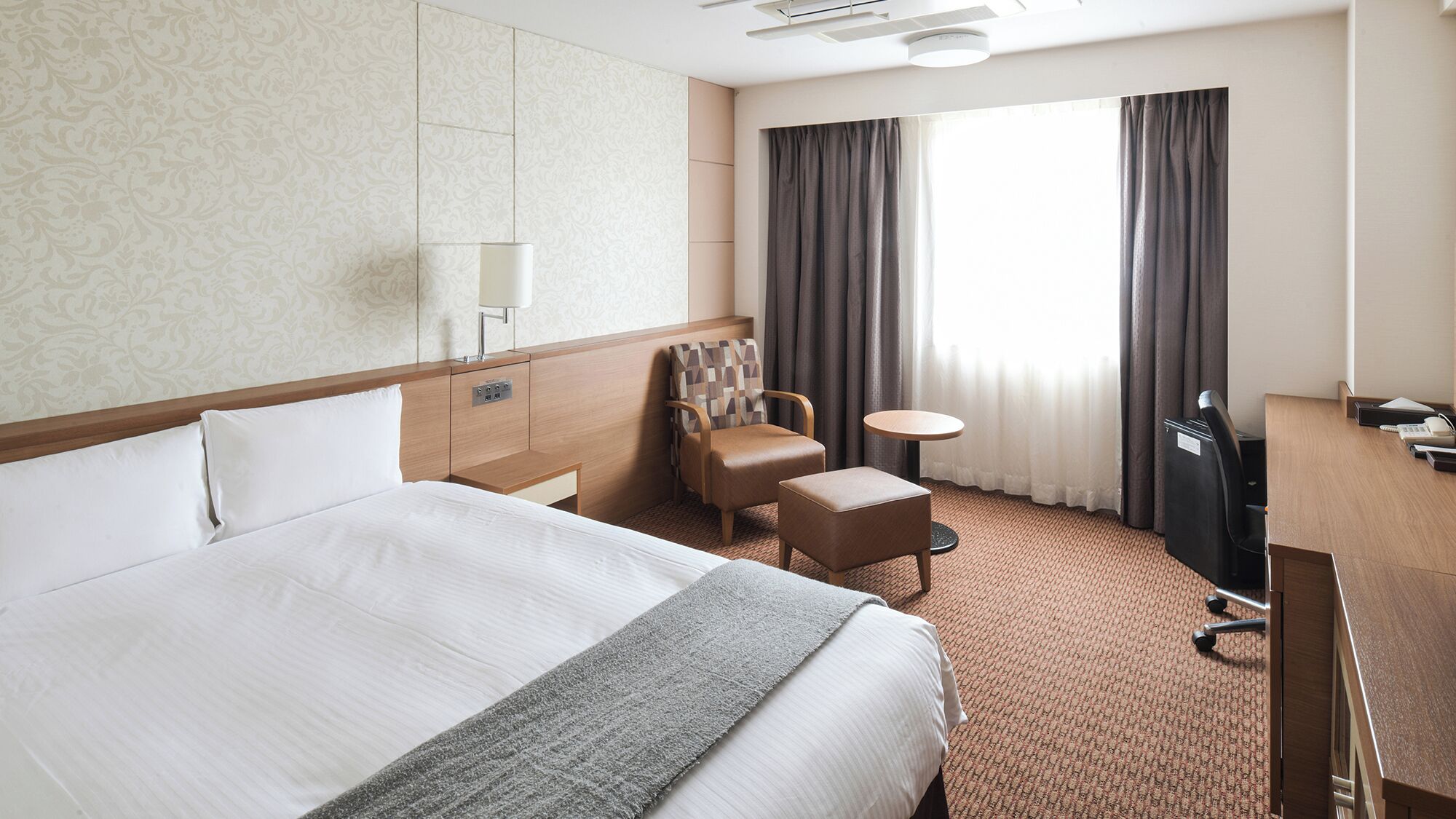 A hotel room with a large bed, a desk, a chair, a small table, and a window with curtains.