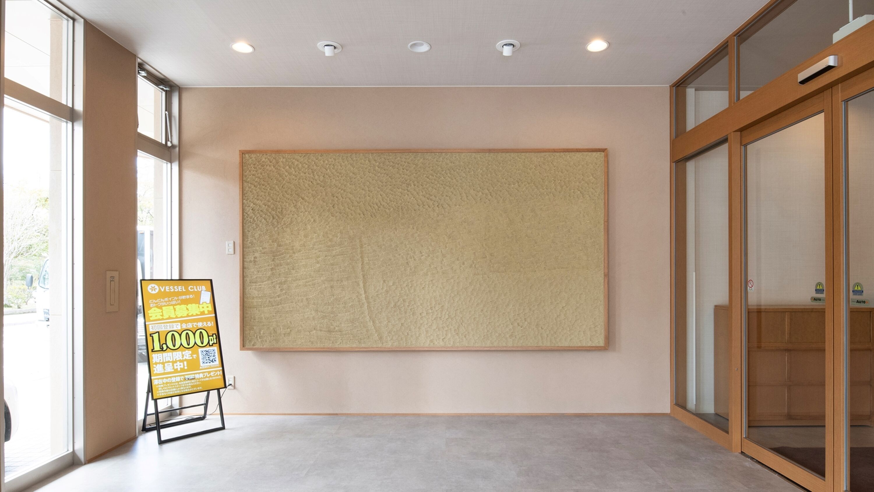 A spacious room with large windows, a wooden floor, and a large textured wall panel.