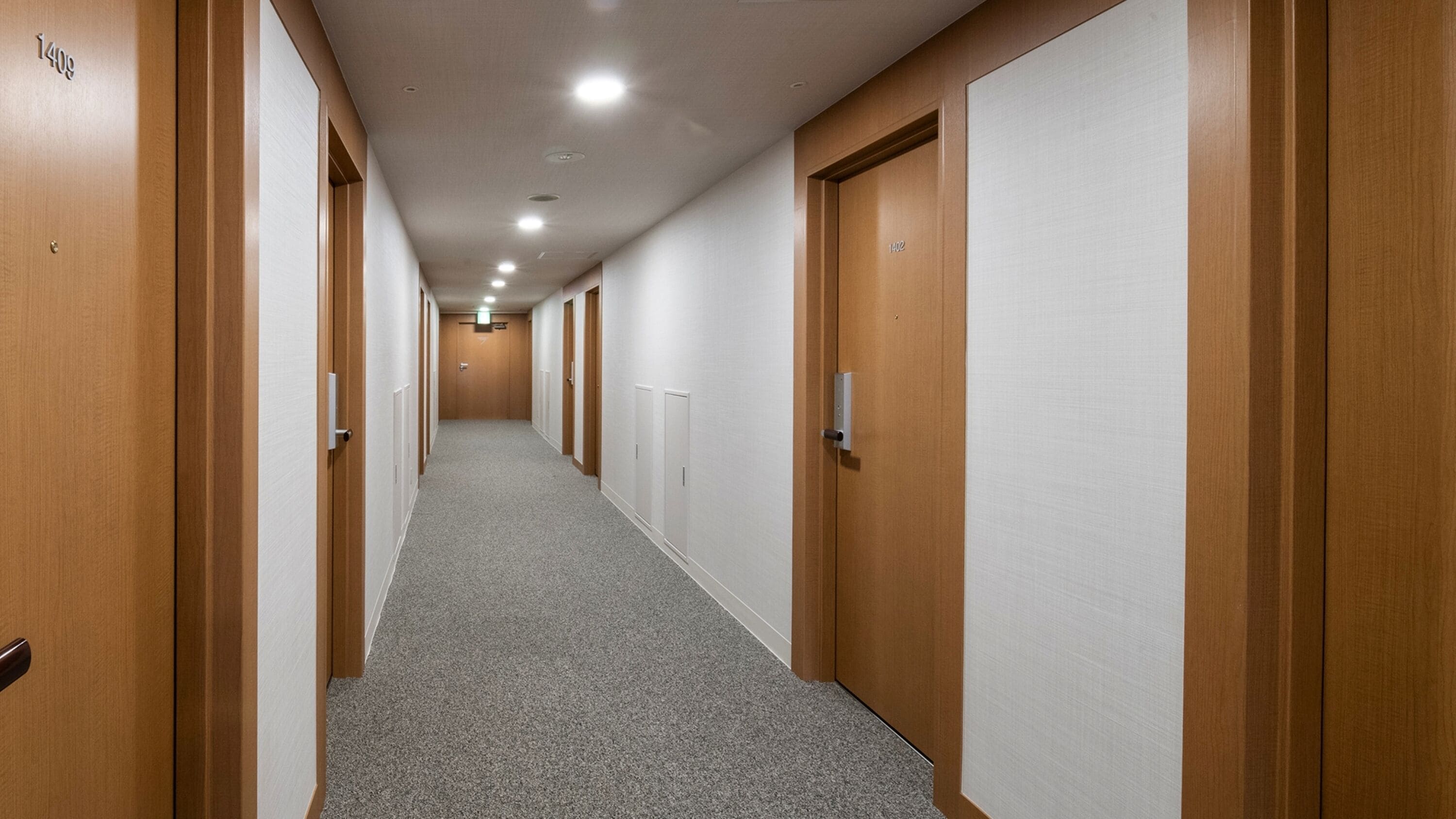 A long, narrow hallway with wooden doors on both sides, carpeted floor, and recessed ceiling lights.