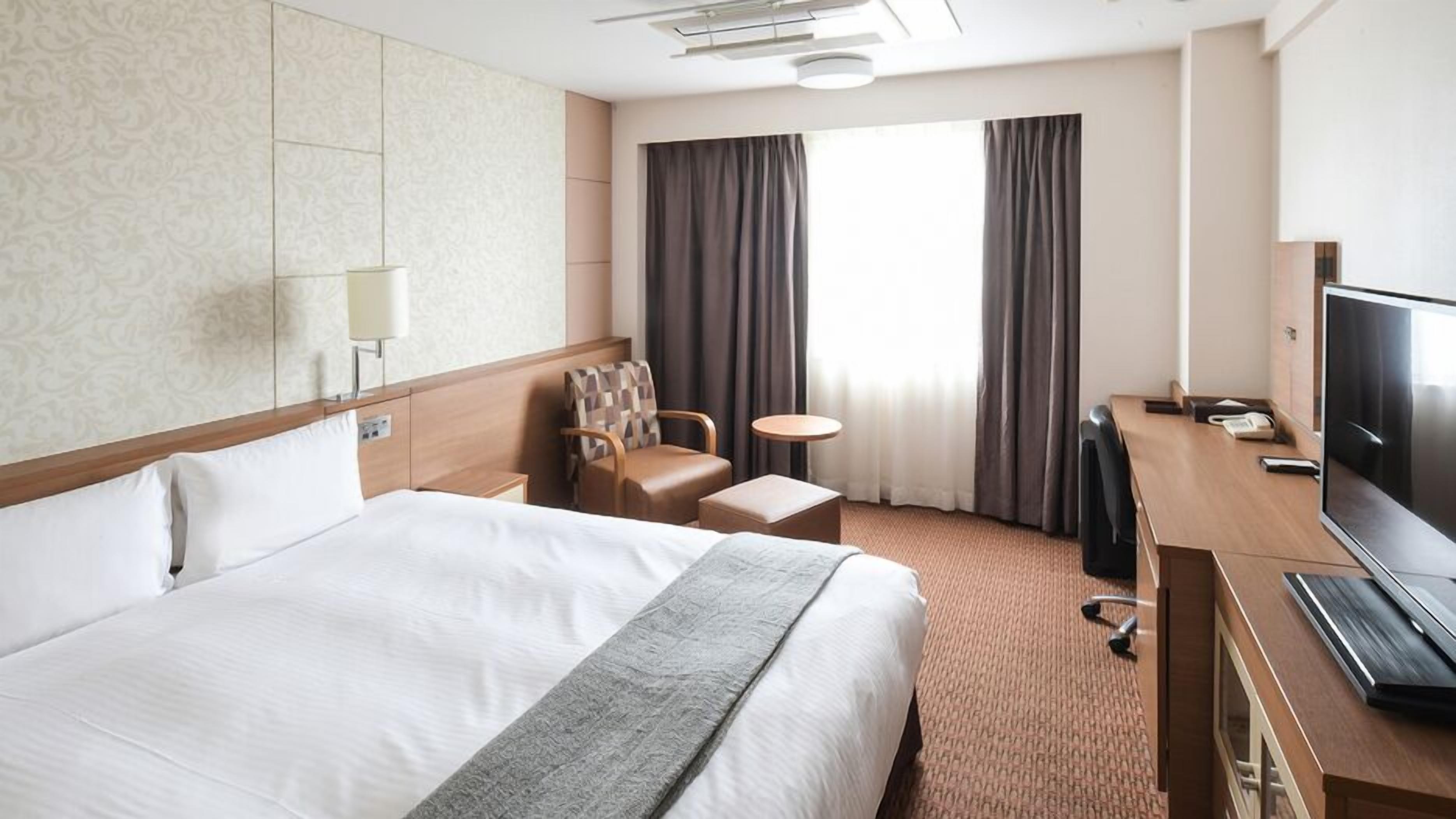 A hotel room with a large bed, a desk with a TV, a chair, a small table, and a window with curtains.
