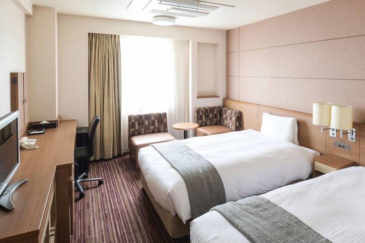 Ein Hotelzimmer mit zwei Betten, einem Schreibtisch mit Computer, einem Stuhl, einem kleinen Tisch und einem Fenster mit Vorhängen.