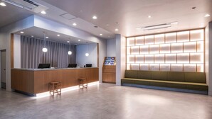 A modern reception area with a wooden reception desk, two bar stools, a seating area with a green sofa, and a wall-mounted display shelf. - Vessel Inn Fukuyama Station (Fukuyama)