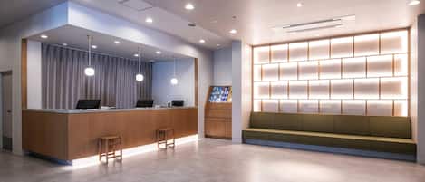 A modern reception area with a wooden reception desk, two bar stools, a seating area with a green sofa, and a wall-mounted display shelf.