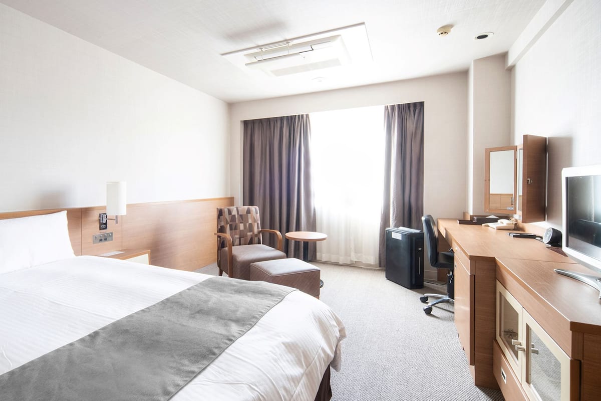 Ein Hotelzimmer mit einem großen Bett, einem Schreibtisch mit Computer, einem Stuhl, einem kleinen Tisch und einem Fernseher.