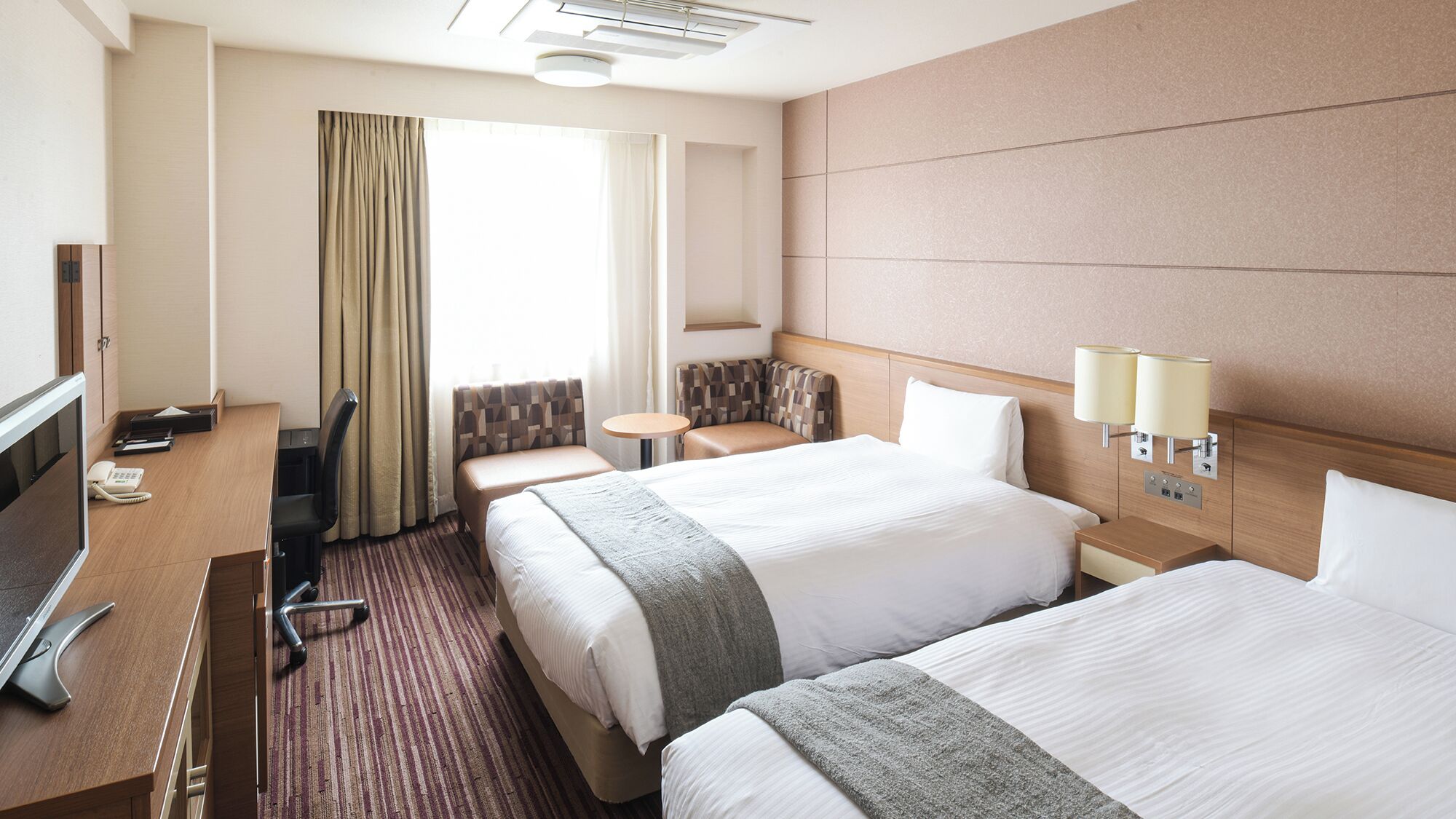 A hotel room with two beds, a desk with a telephone, a chair, a small table, and a window with curtains.