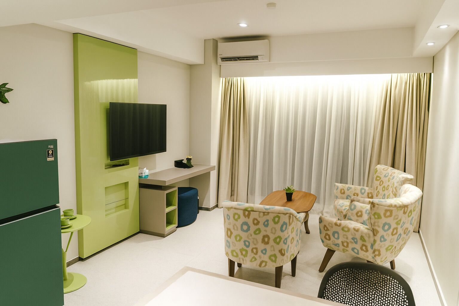 A compact hotel room with a green minibar, a flat-screen TV, a small table with a plant, two patterned armchairs, and a large window with curtains.
