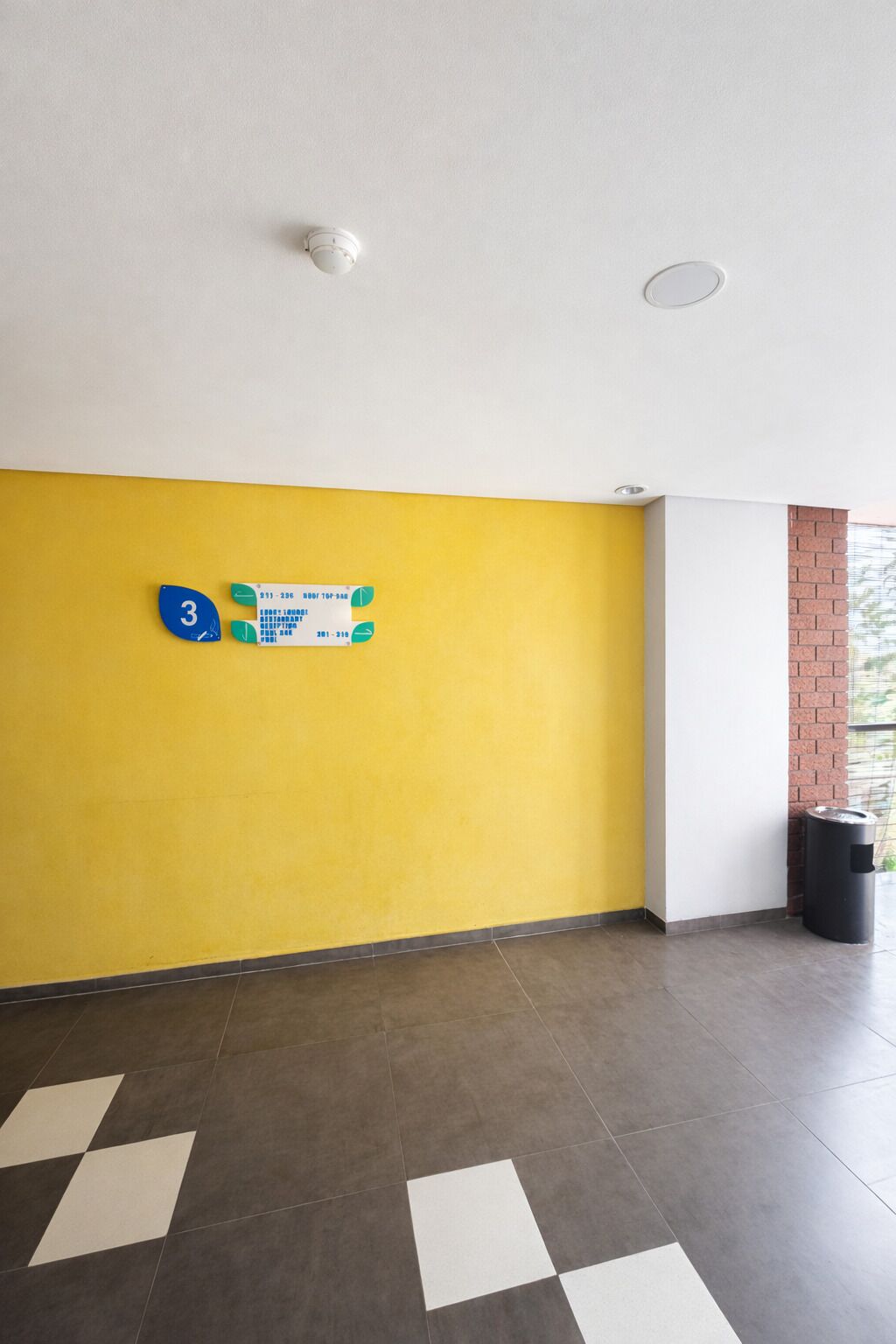 A hallway with a yellow wall, a white wall, and a grey tiled floor. There is a trash can and a number sign on the wall.