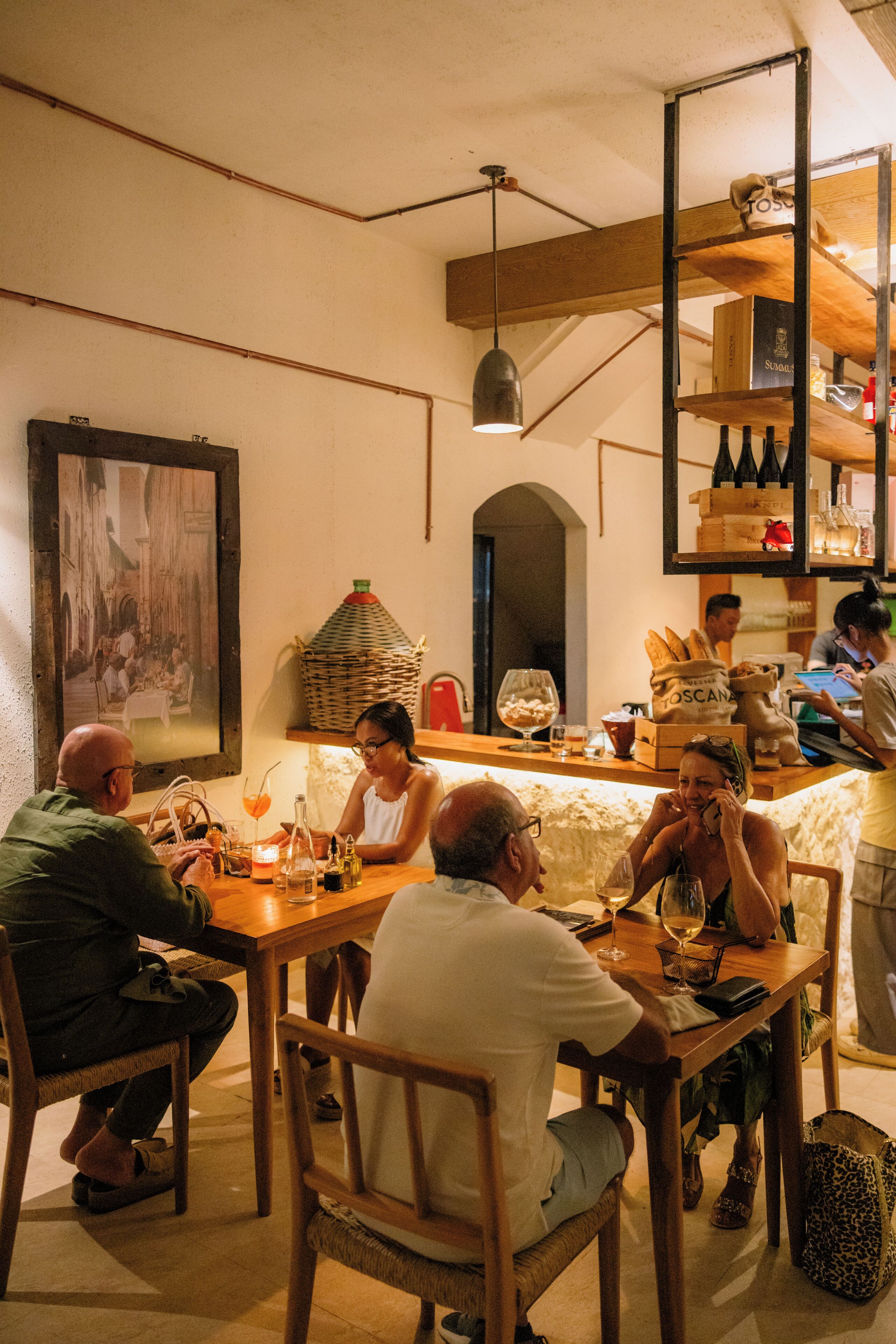 A cozy restaurant interior with wooden tables and chairs, a hanging light fixture, and a wall-mounted shelf with bottles.
