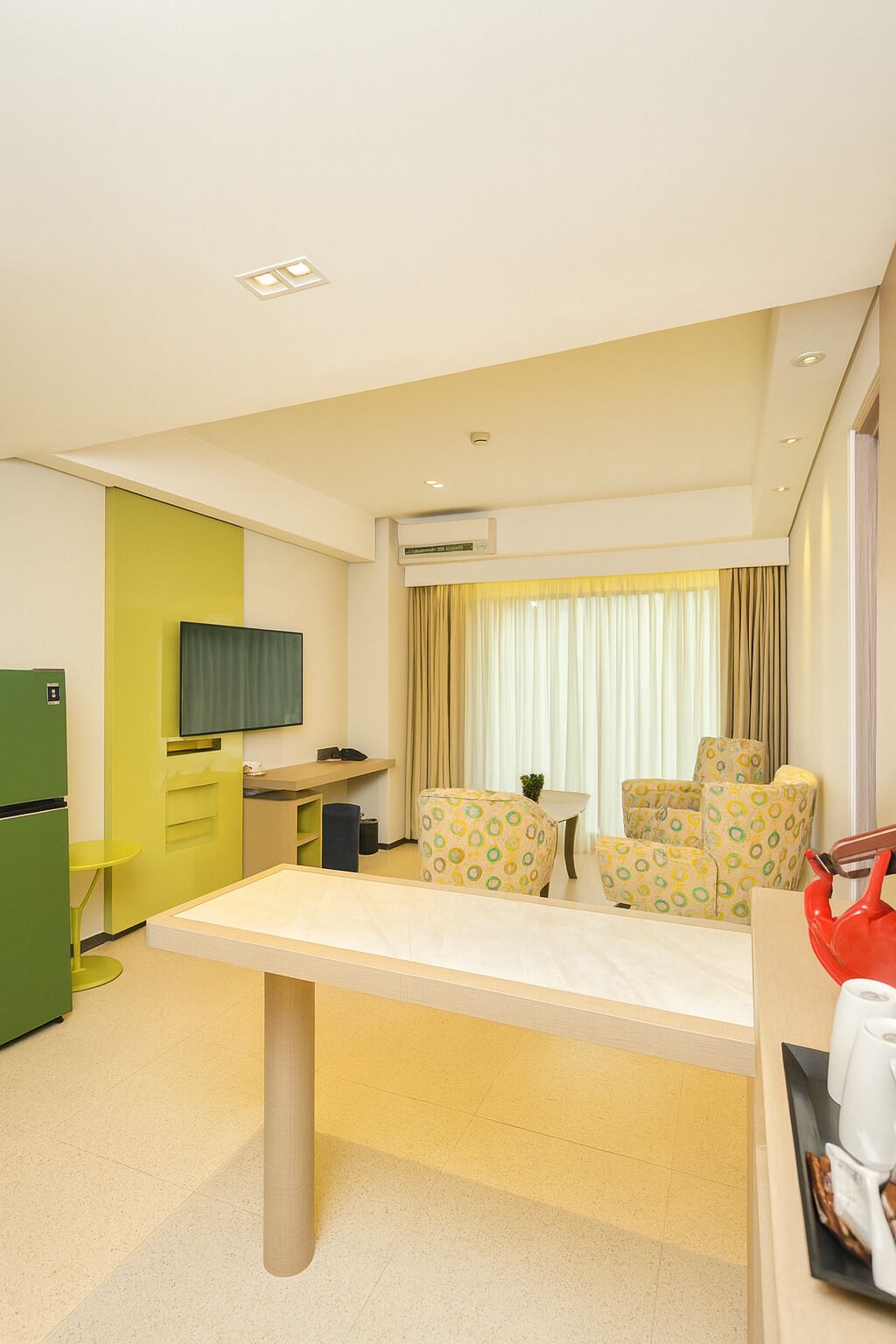 A hotel room with a dining table, four patterned chairs, a green refrigerator, a television, and a window with curtains.