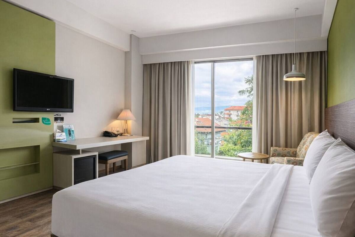 A hotel room with a large bed, a desk with a lamp, a television, and a view of the city.