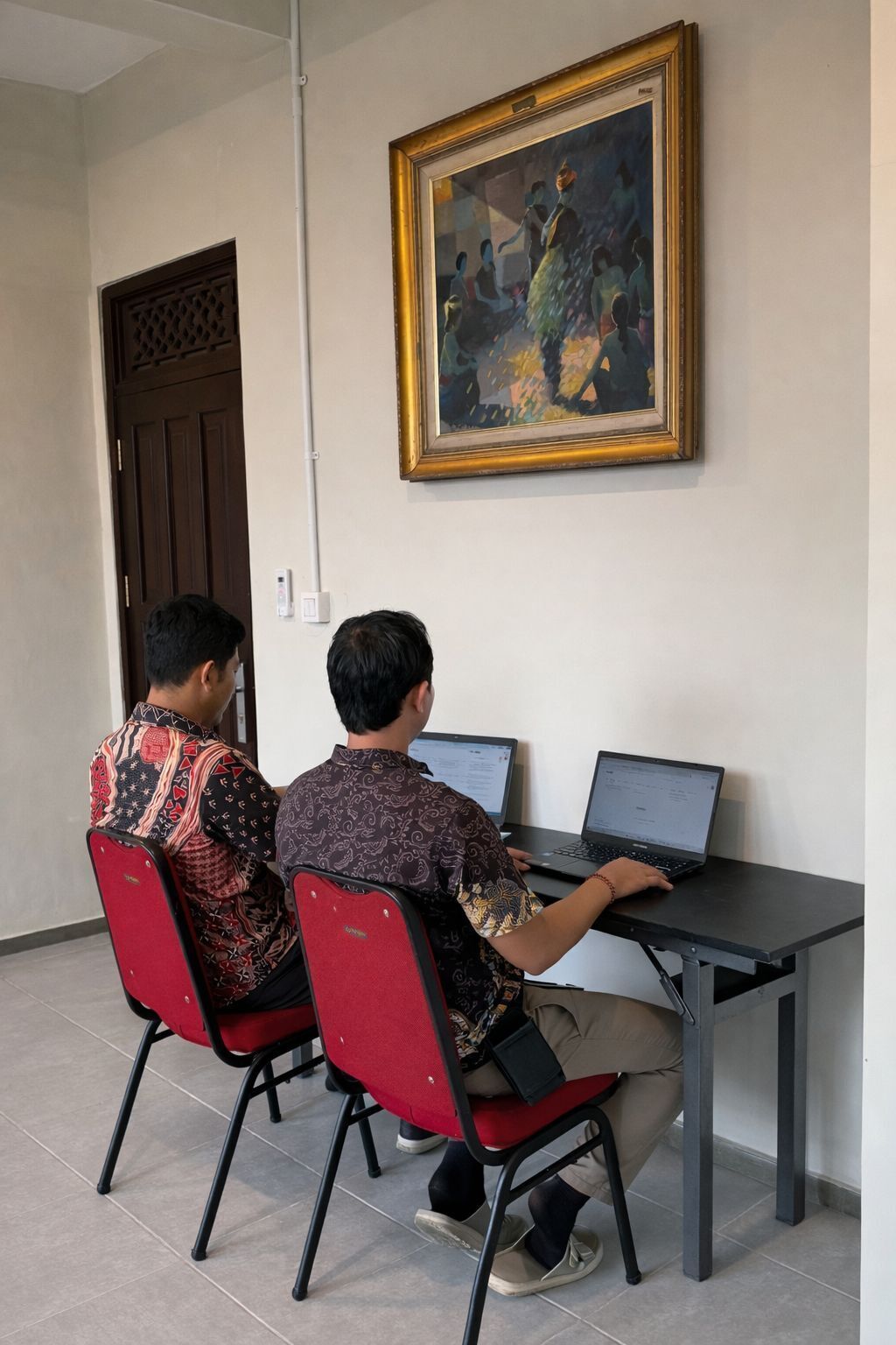 Two individuals seated at a table with laptops, in a room with a door and a framed painting on the wall.