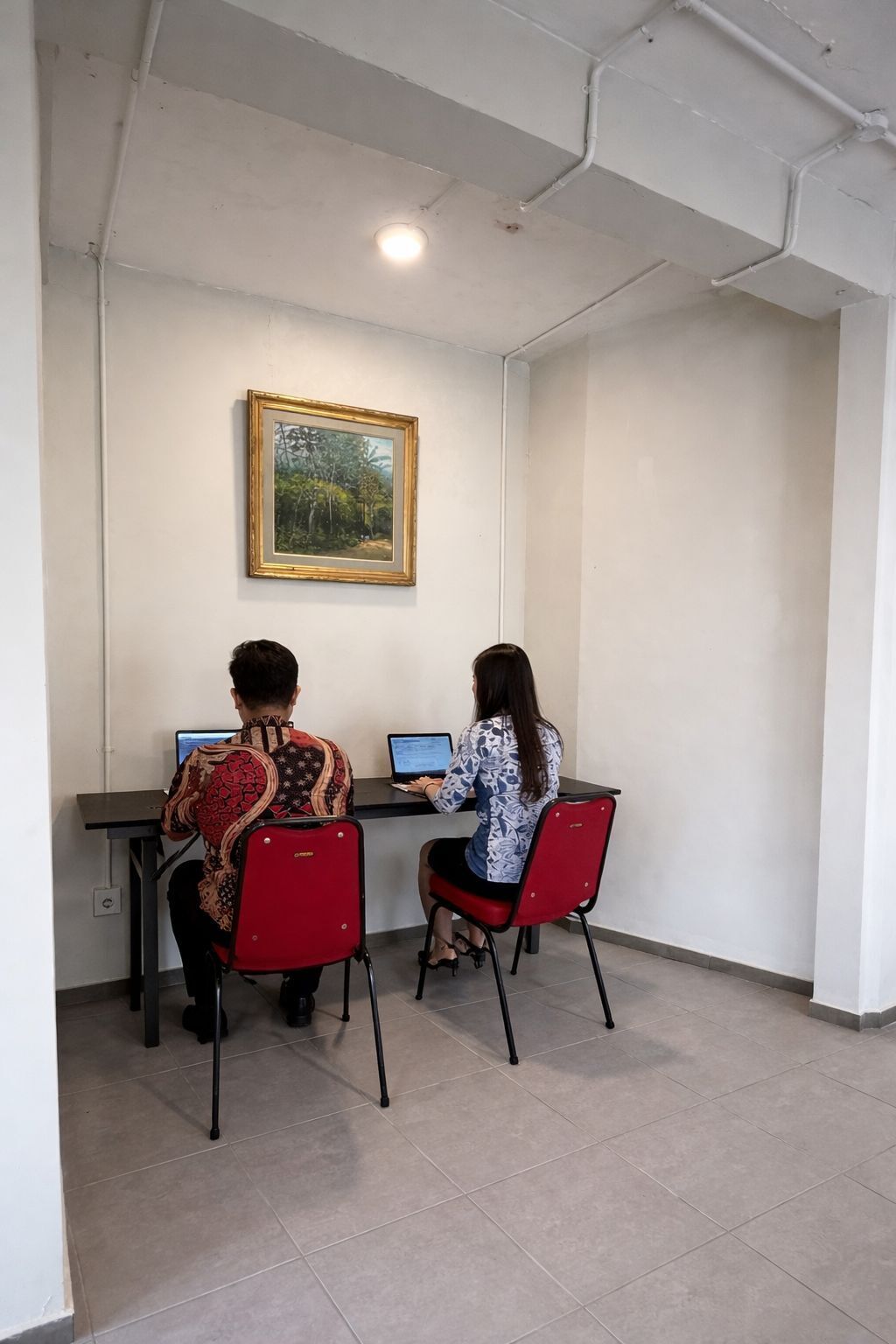 Two people working at desks in a room with tiled flooring, a painting on the wall, and a ceiling with exposed pipes.