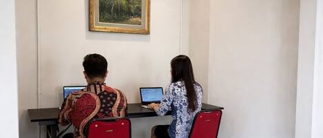 Two people working at desks in a room with tiled flooring, a painting on the wall, and a ceiling with exposed pipes.