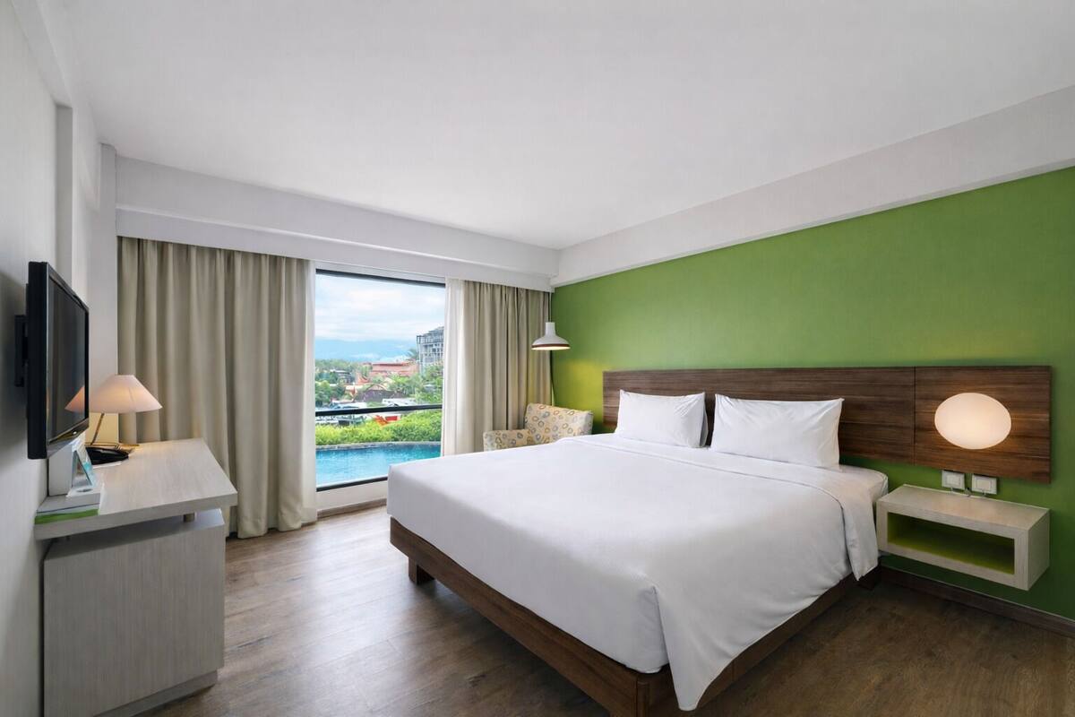 Habitación de hotel con una cama grande, un escritorio con televisor, una lámpara y vistas a una piscina y edificios.
