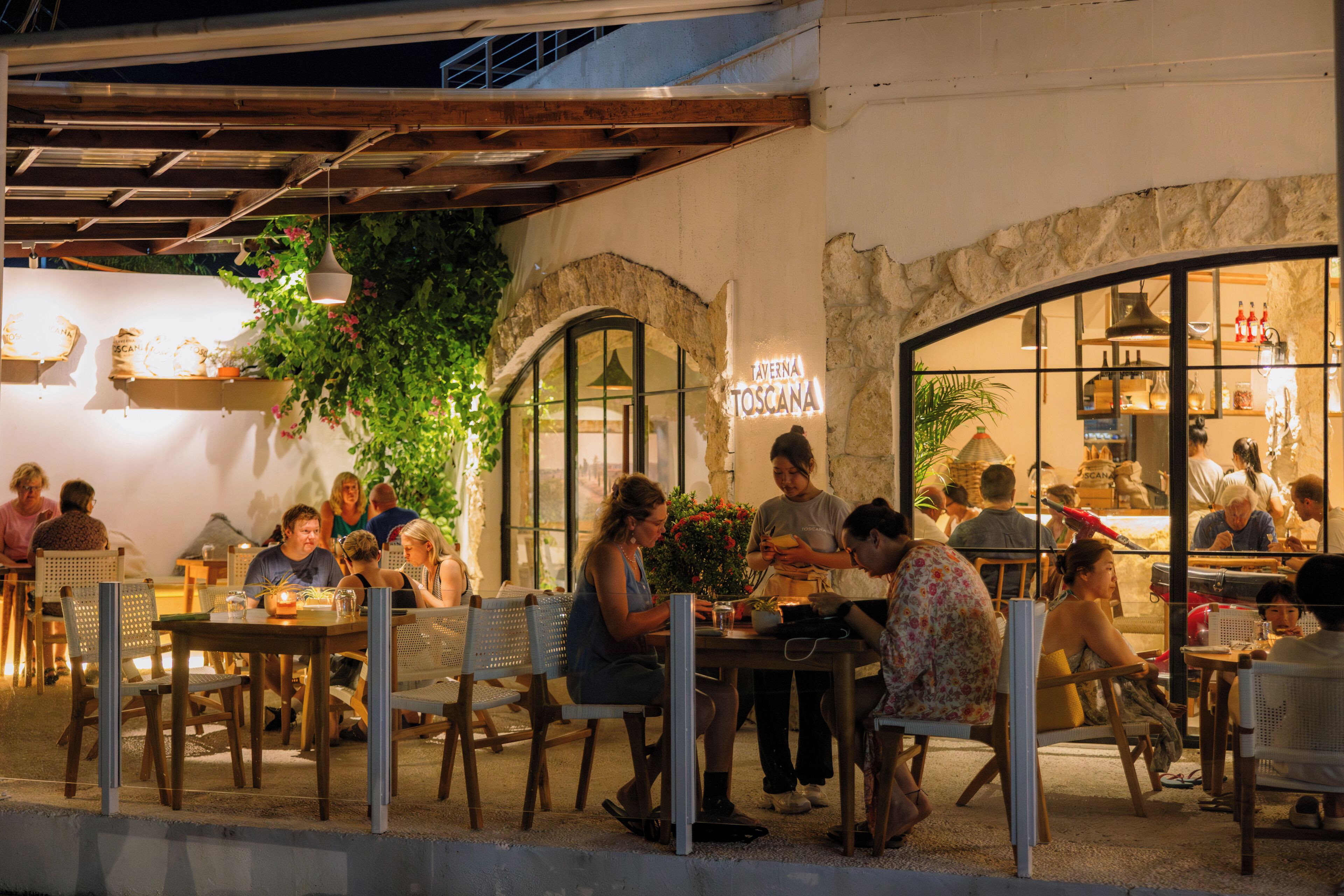 An outdoor dining area with tables and chairs, a stone wall, and a sign reading "Taverna Toscana".