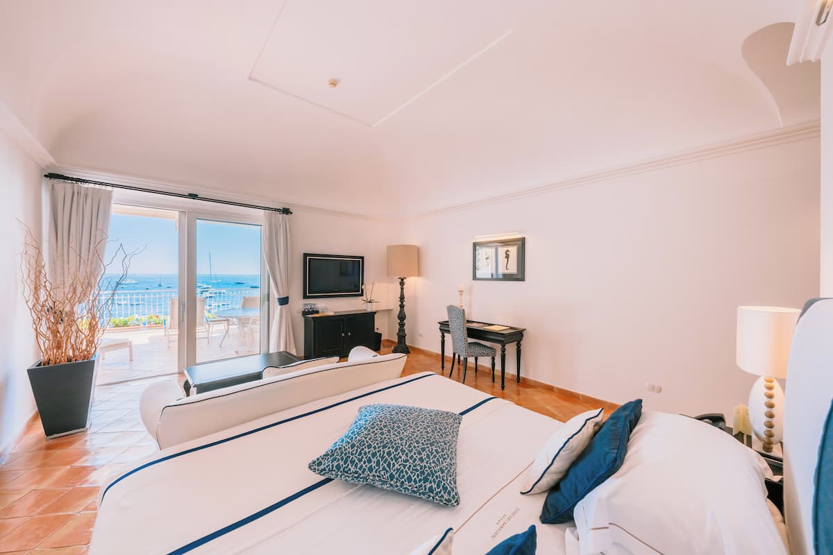 A hotel room with a bed, a desk, a chair, a TV, and a view of the sea.