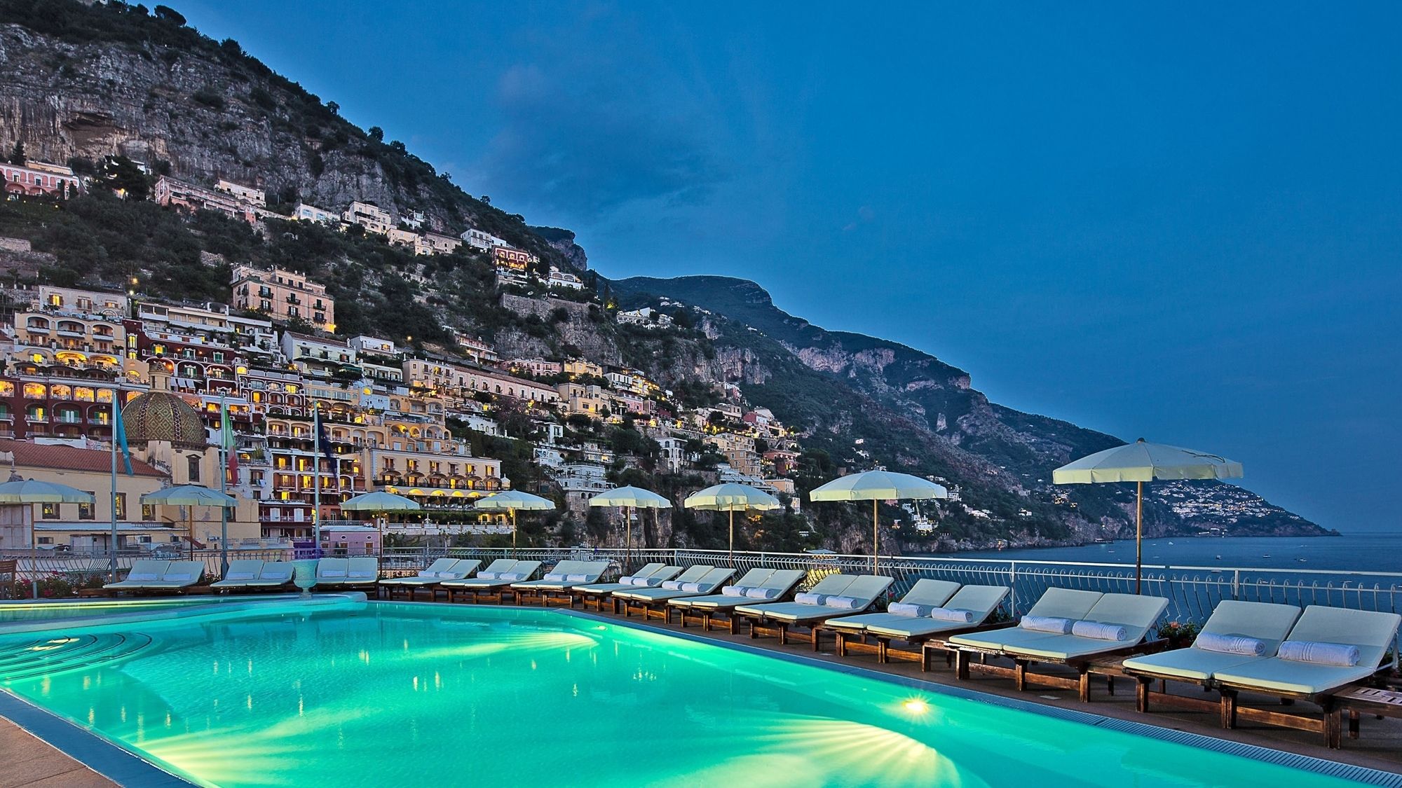 A swimming pool with lounge chairs and umbrellas, set against a backdrop of terraced buildings and a mountainous coastline.