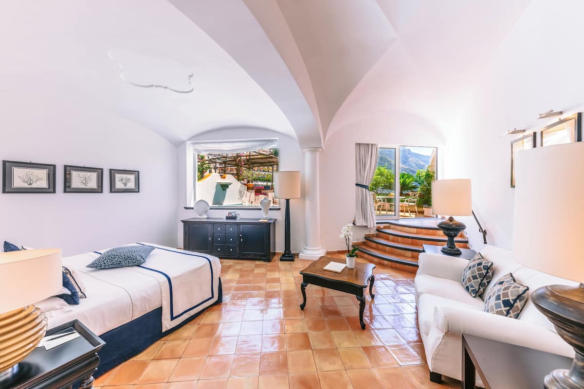 A spacious hotel room with a bed, a sofa, a coffee table, and a view of a terrace with plants and a pool.