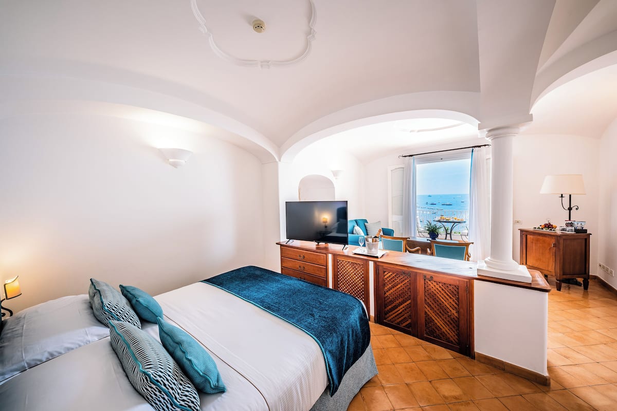 A hotel room with a bed, a desk, a television, and a view of the sea.