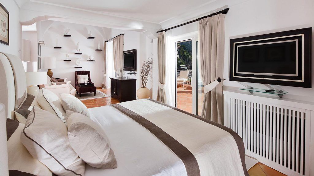 A spacious bedroom with a large bed, a TV mounted on the wall, and a view of the outdoors through a sliding glass door.