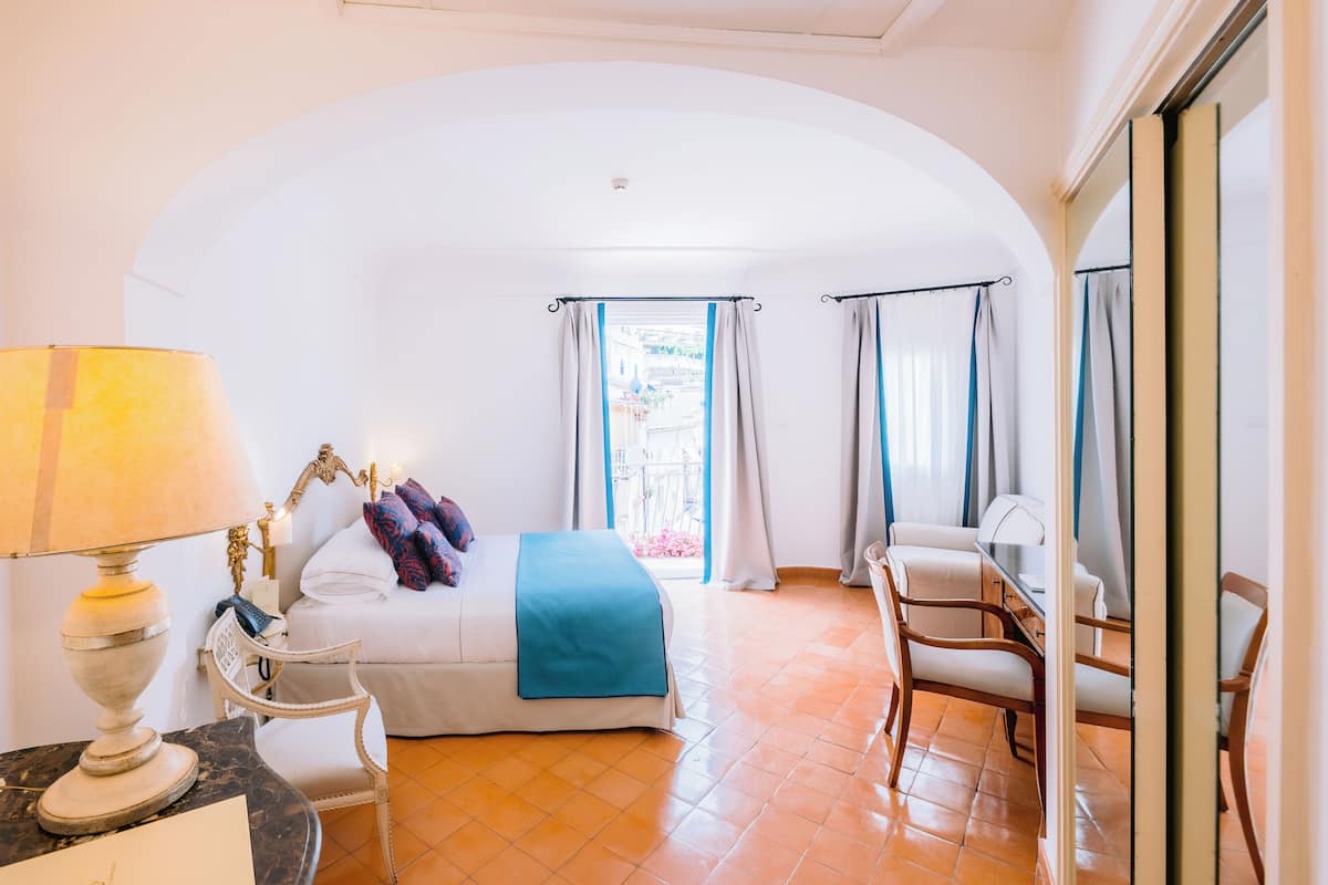 A hotel room with a bed, a desk, a chair, a lamp, and a view of the outside.