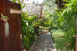 Ein hölzernes Tor mit einem 'Welcome'-Schild, ein reetgedecktes Gebäude und ein gepflasterter Weg, umgeben von üppiger Vegetation.