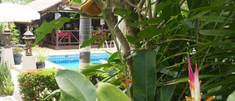 A pool area with a wooden deck, surrounded by lush greenery and a building with a red roof.