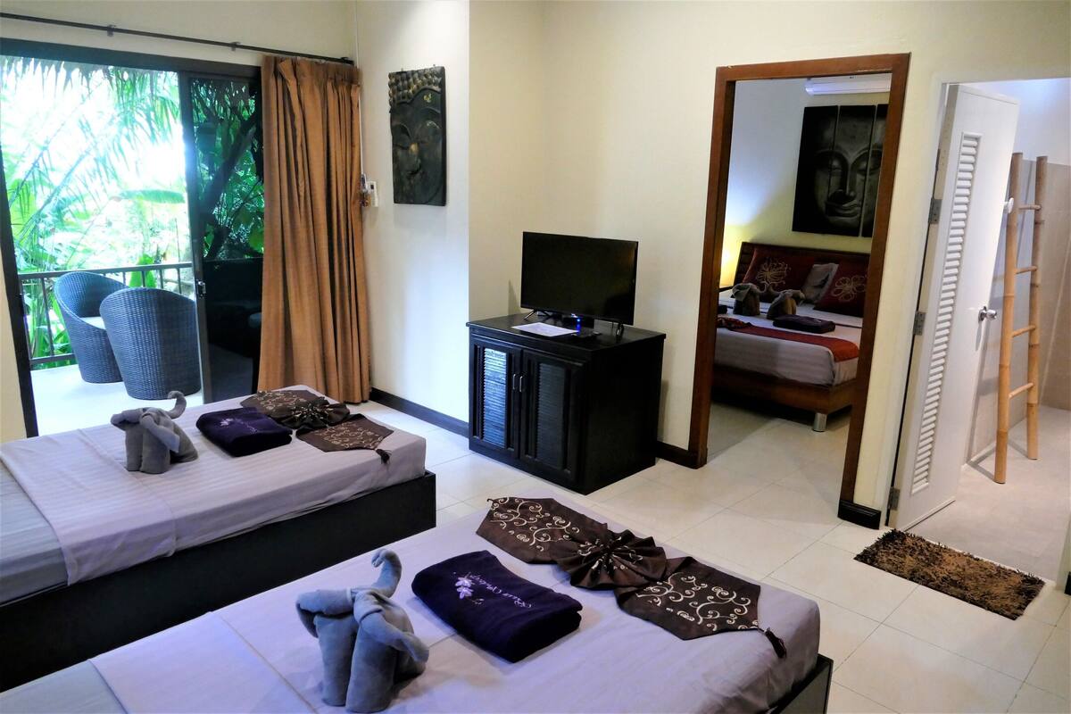 A hotel room with two beds, a TV, a balcony with wicker chairs, and a view of greenery.