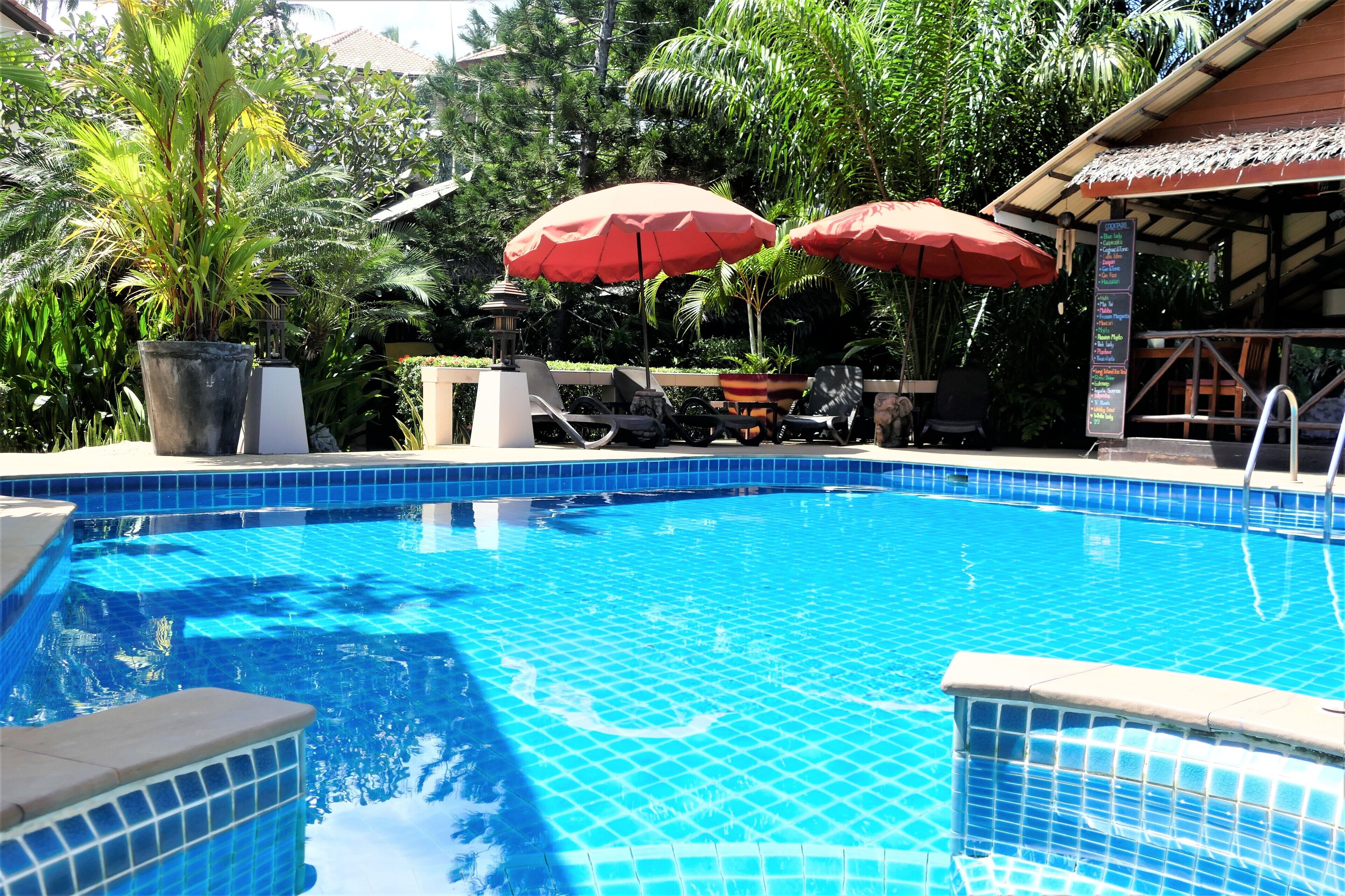 Piscina con baldosas azules, rodeada de vegetación y sombrillas rojas.