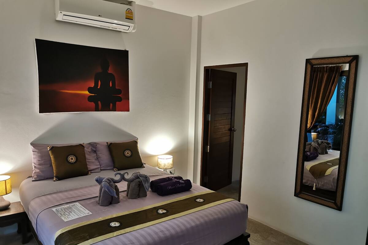 A neatly arranged bedroom with a bed, bedside lamps, a wall-mounted air conditioner, a door, and a mirror reflecting the room.