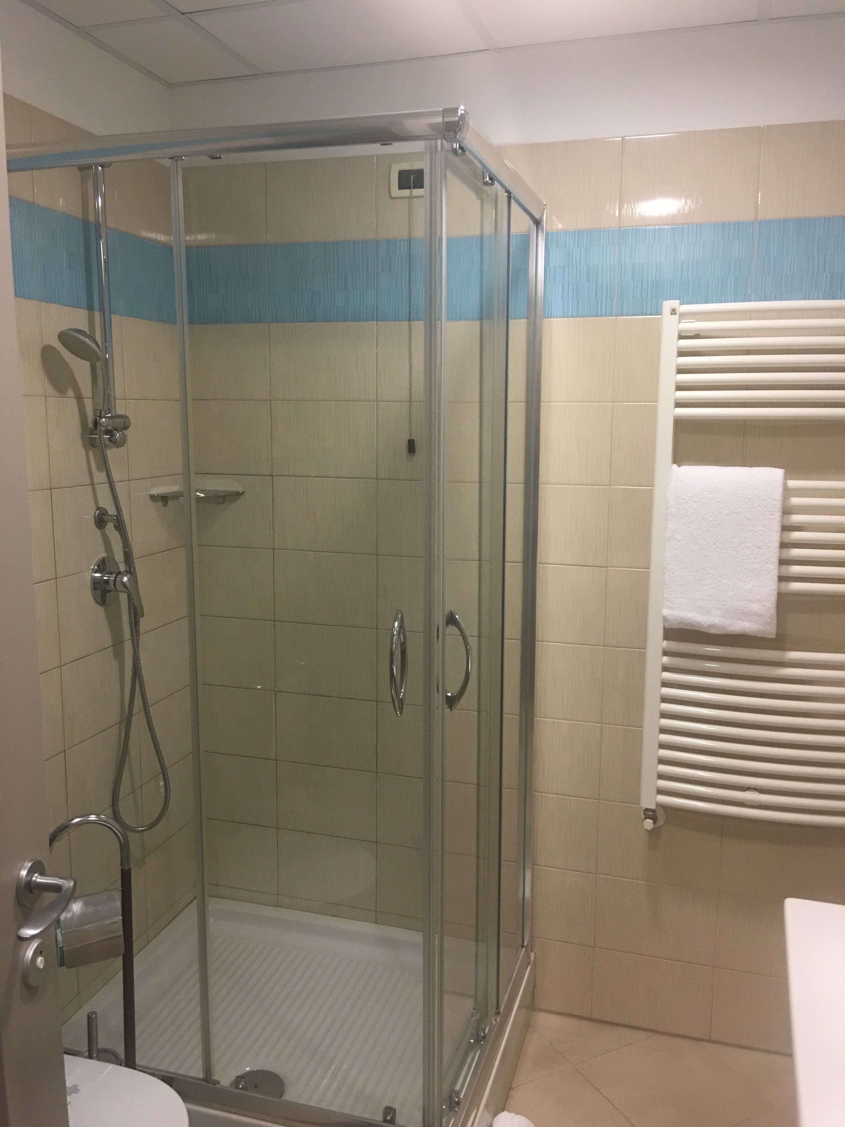 Una cabina doccia in vetro con soffione a mano, un porta asciugamani con un asciugamano bianco e una parete rivestita di piastrelle con una striscia blu.