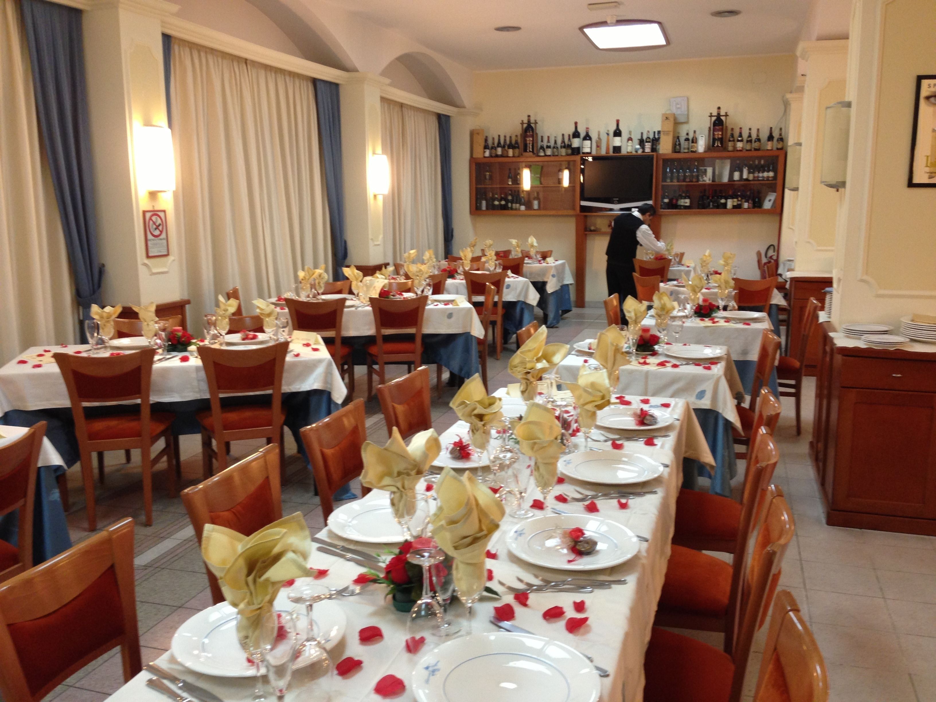A formal dining area with tables set for multiple guests, a bar in the background, and a person standing near the bar.
