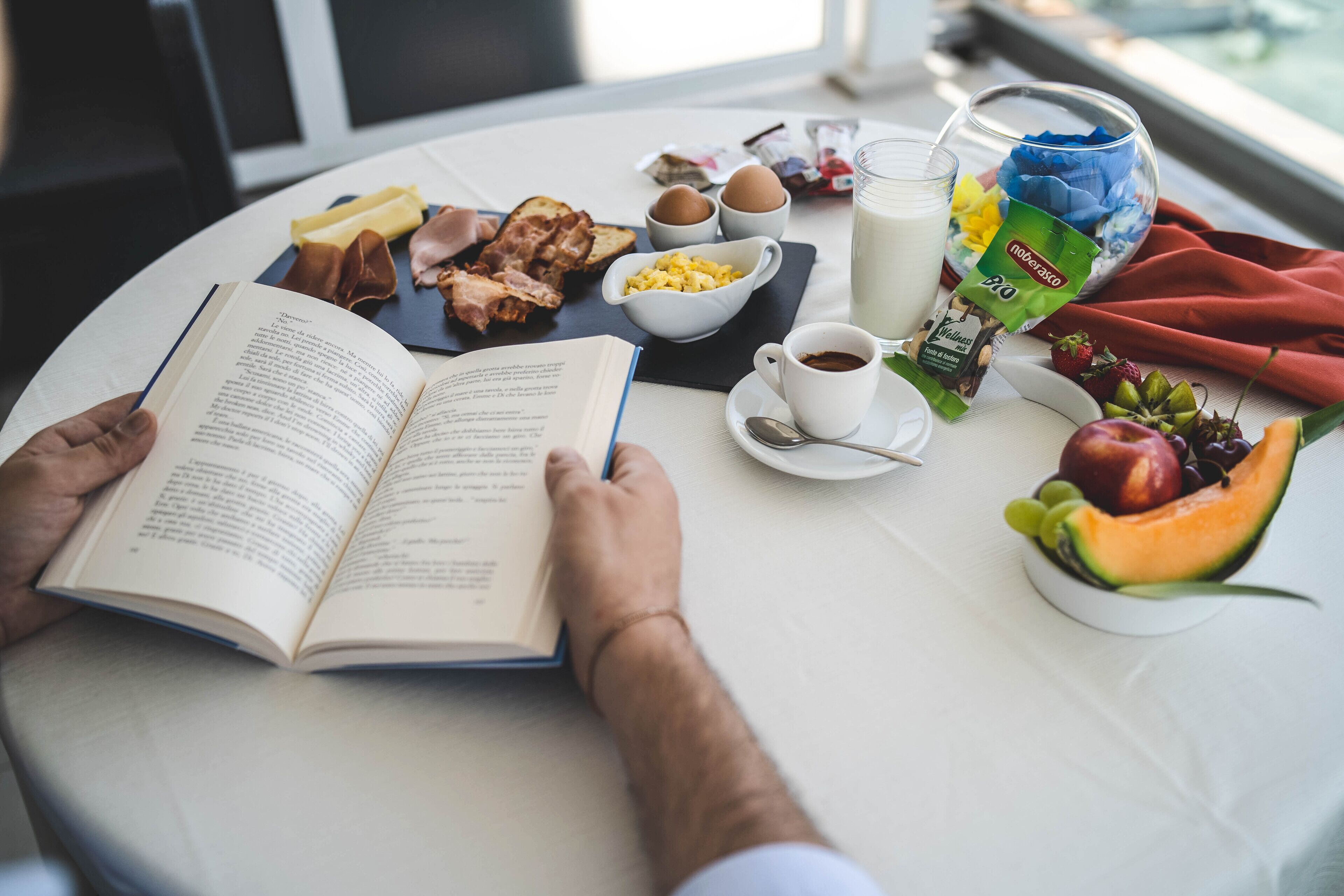 Una persona leyendo un libro en una mesa con alimentos para el desayuno, como tostadas, huevos, leche, café y fruta.
