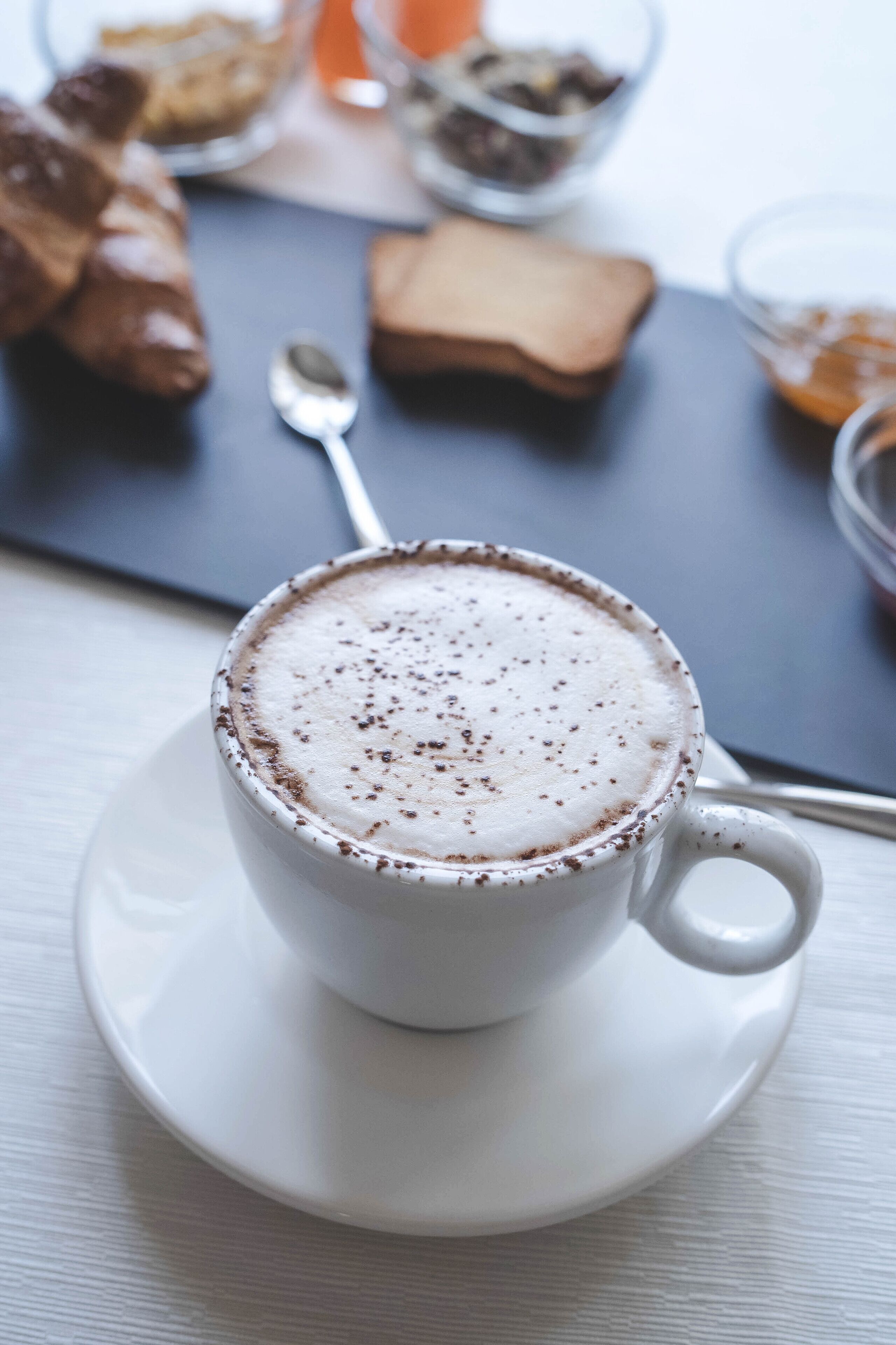 A cup of coffee with a frothy top, a spoon, and a pastry on a table.