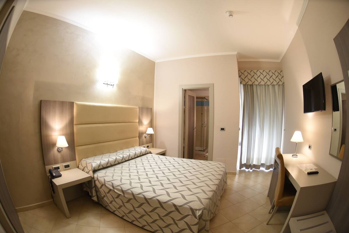 A hotel room with a bed, bedside tables, a desk, a chair, a TV, and a bathroom door.