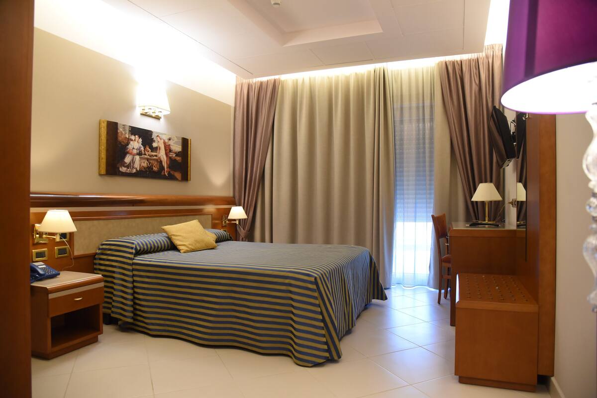 A hotel room with a bed, bedside tables, a desk, a chair, and a large window with curtains.