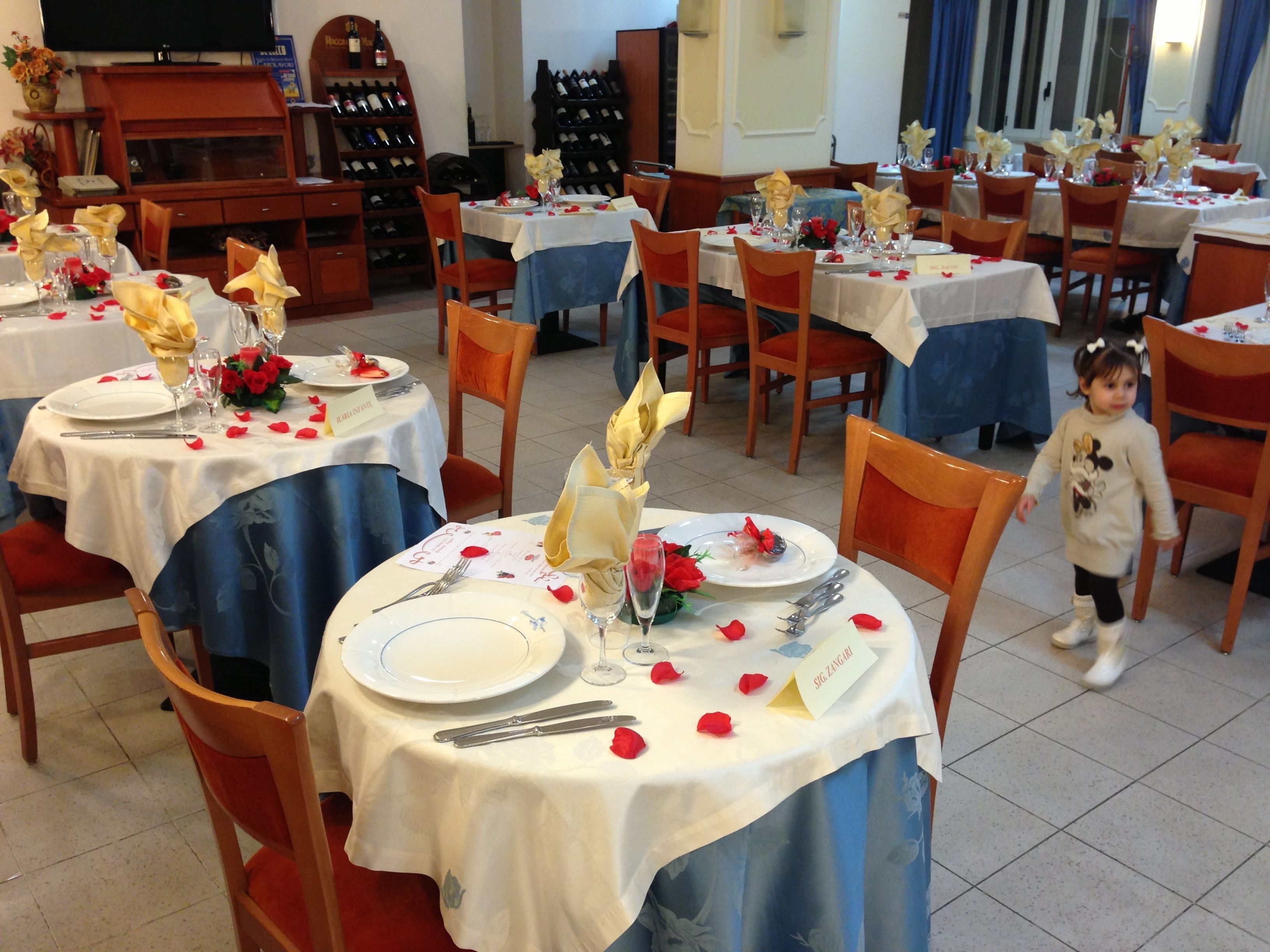 A restaurant with round tables set for dining, each with a white tablecloth, red rose petals, and a wine glass.