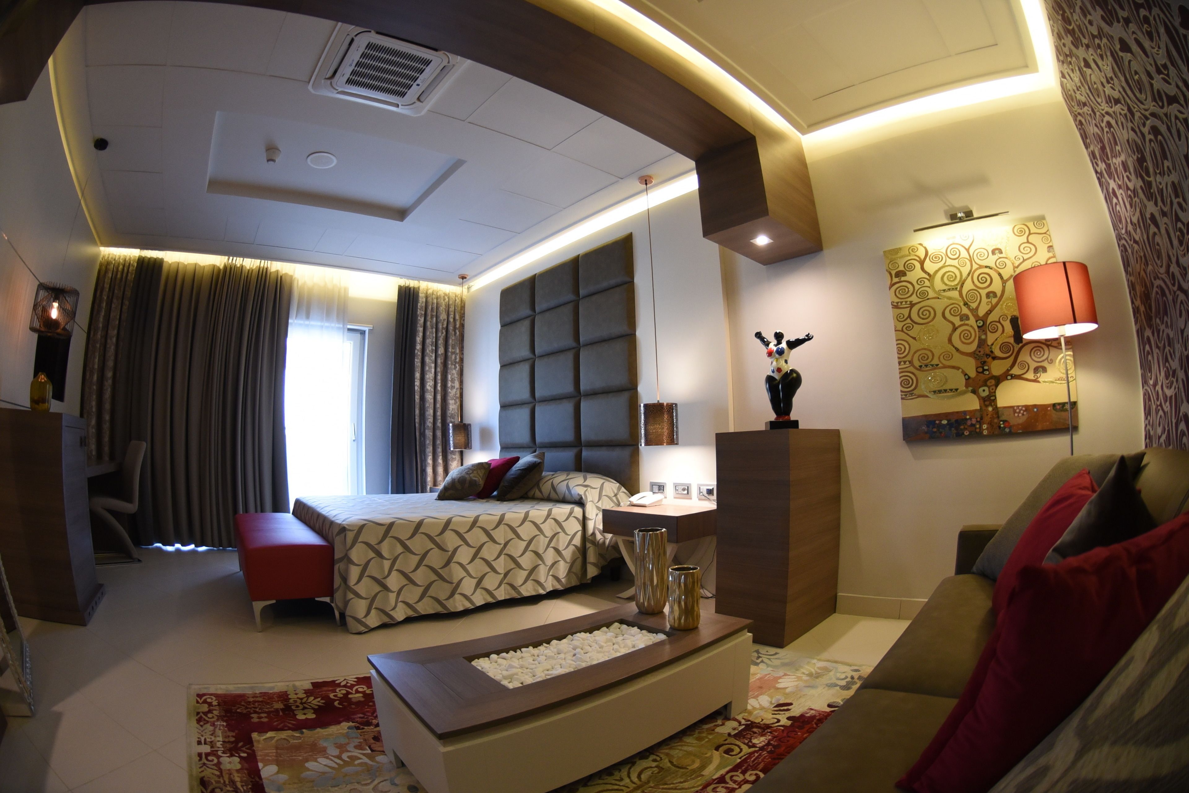 Una habitación de hotel moderna con una cama grande, un área de descanso con sofá y una mesa de centro, un escritorio y un ventanal con cortinas.