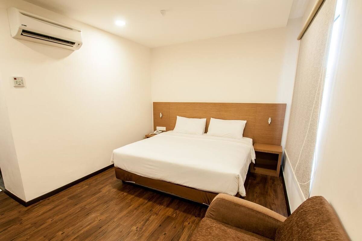 Quarto de hotel com piso de madeira, cama com roupa de cama branca, cabeceira de madeira, mesa de cabeceira e poltrona marrom.