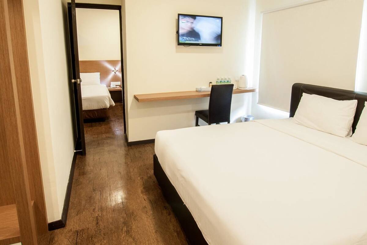 Quarto de hotel com uma cama grande, uma escrivaninha com cadeira, uma televisão na parede e chão de madeira.